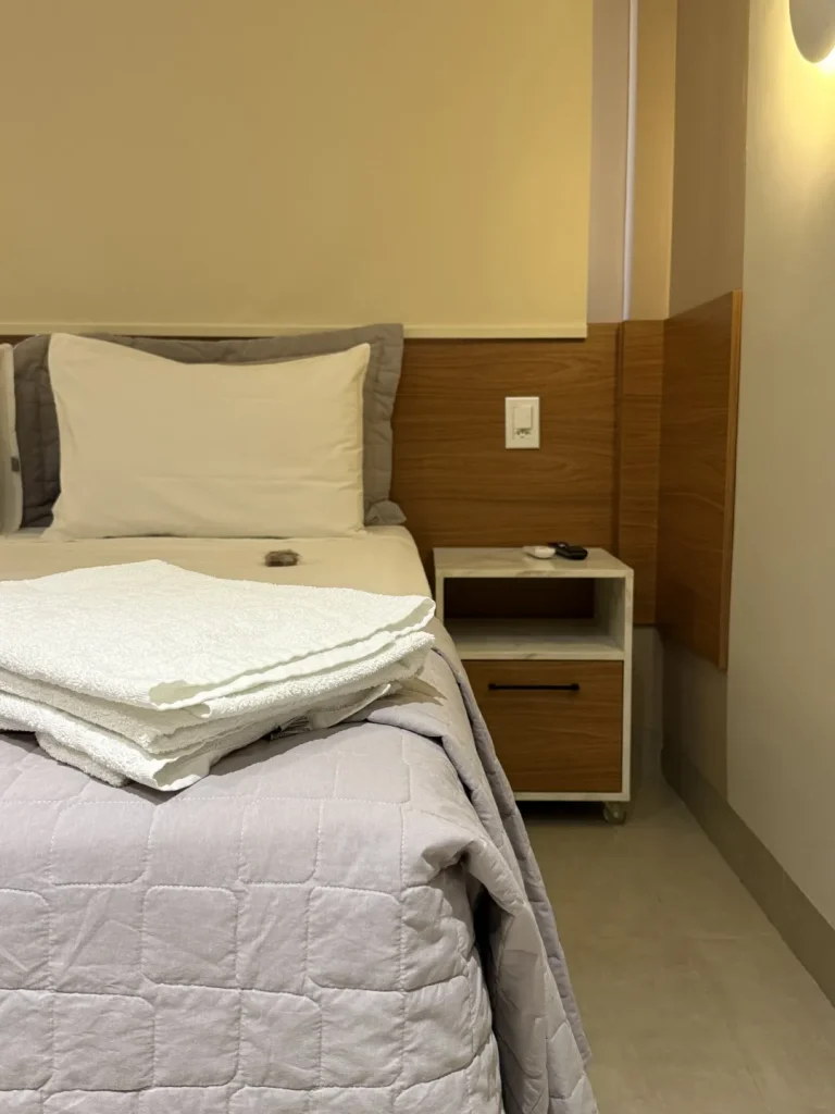 Detalhe da cama queen com enxoval dobrado e criado-mudo ao lado
