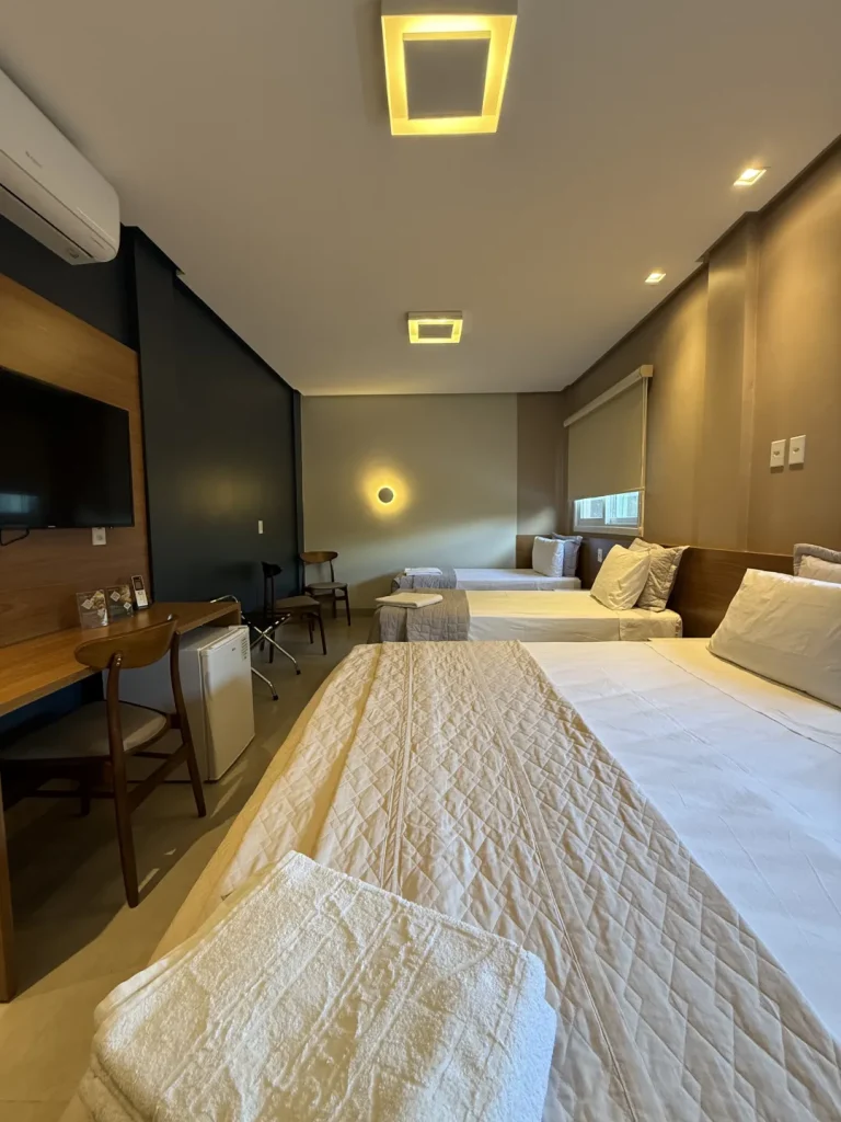 Cama queen em primeiro plano, com as camas de solteiro e a TV ao fundo
