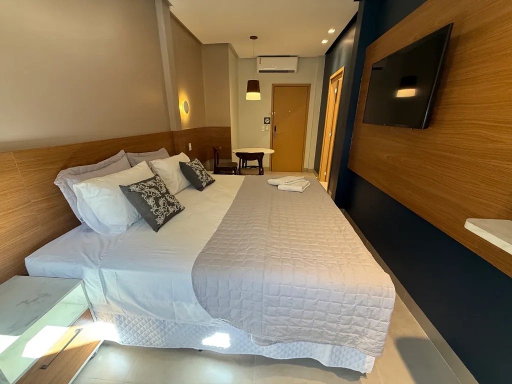 Cama queen vista de lado, com Smart TV no painel escuro