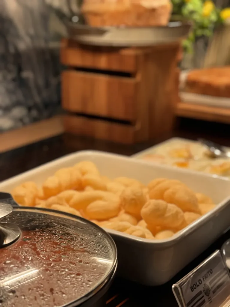 Travessa de bolo frito no buffet quente, com cesta de pães ao fundo.