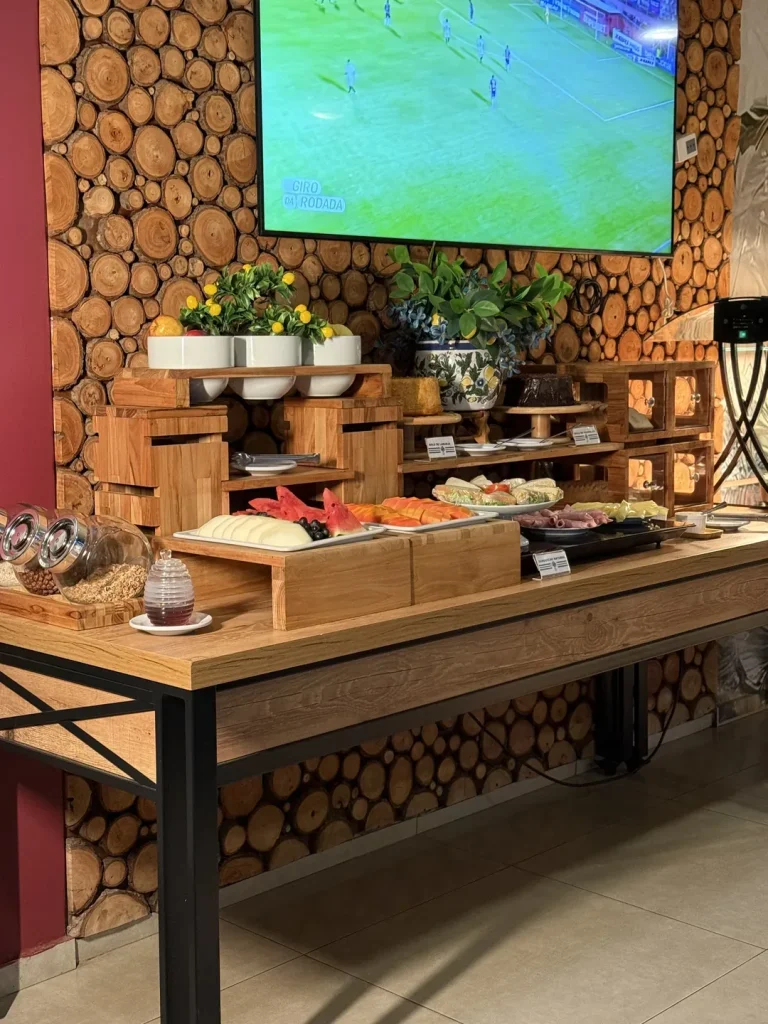 Buffet do café da manhã montado ao longo da parede de toras de madeira, com frutas, bolos, frios e cereais.