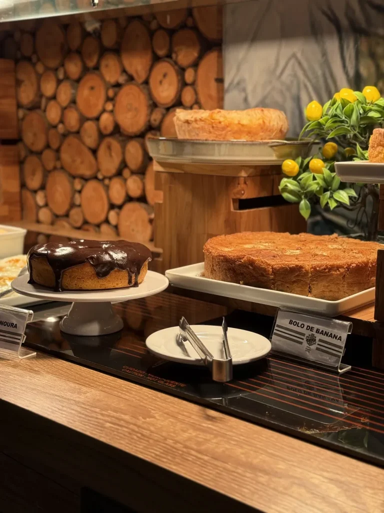 Bolo de cenoura com cobertura de chocolate ao lado do bolo de banana, sobre a bancada aquecida.