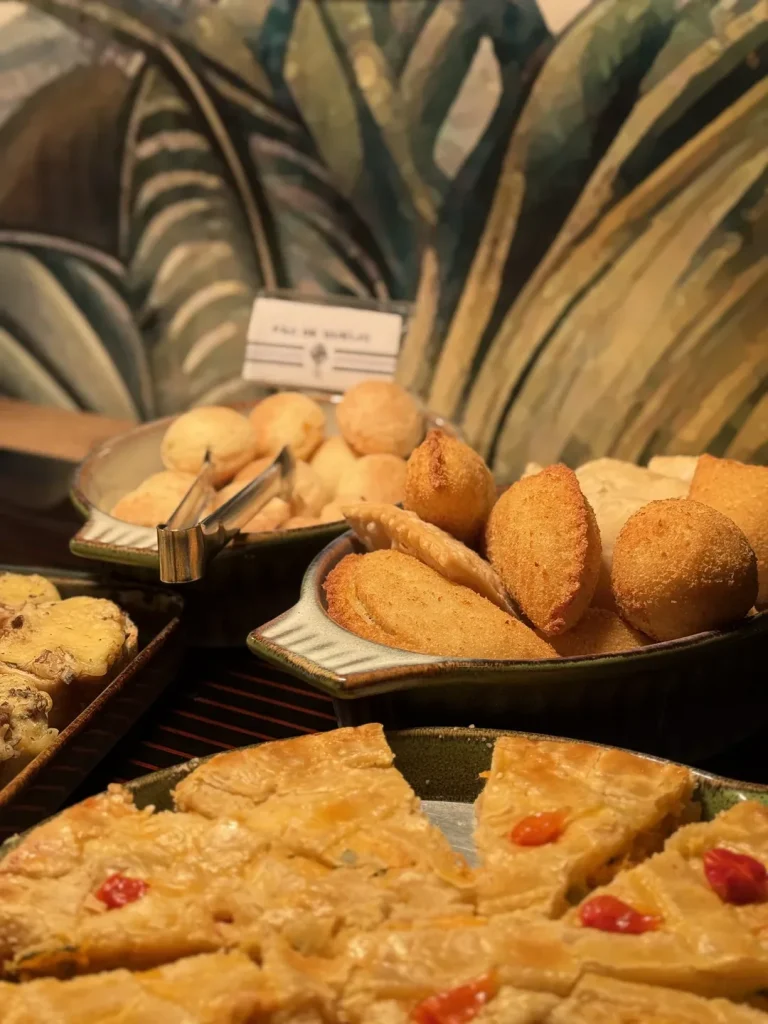 Pão de queijo, risoles e fatias de empadão servidos em travessas de cerâmica.