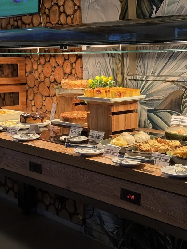Bolos, pão de queijo e empadão expostos sob a proteção de vidro do buffet.