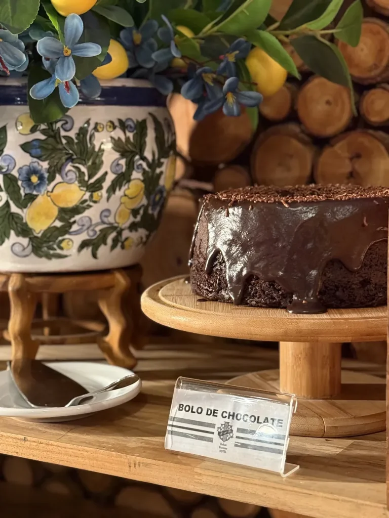 Bolo de chocolate com ganache e granulado, identificado pela placa do buffet.