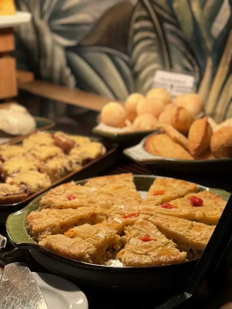 Empadão fatiado em travessa de cerâmica, com pão de queijo e salgados ao fundo.