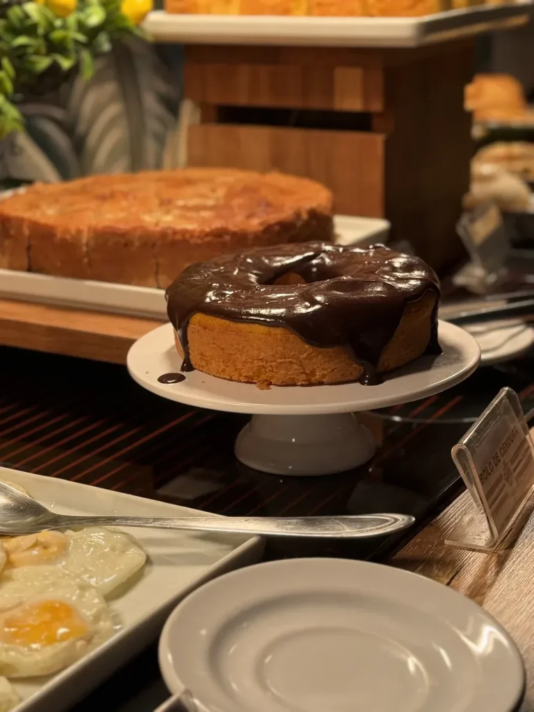 Bolo de cenoura com cobertura de chocolate em suporte de louça, com ovos fritos ao lado.