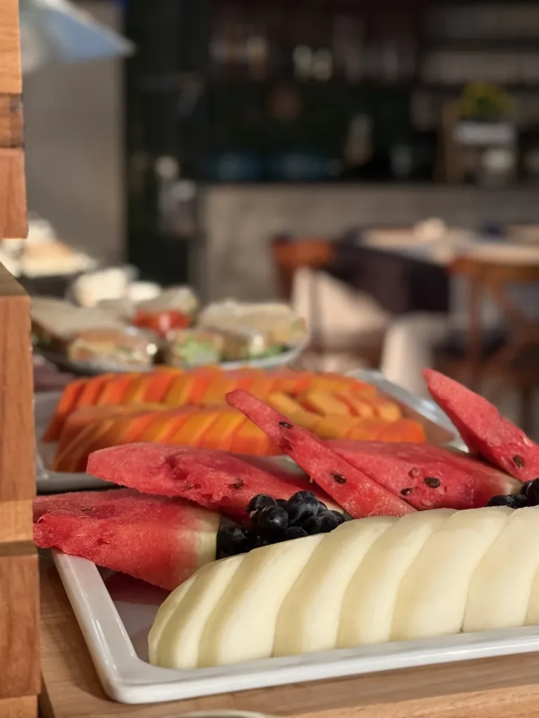 Bandeja com melancia, mamão, melão e uvas, com o salão do restaurante ao fundo.
