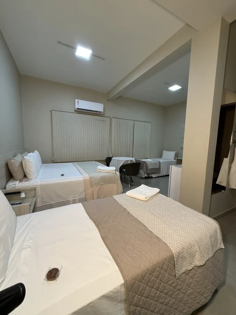 Cama queen em primeiro plano, com mesa redonda e cama de solteiro ao fundo