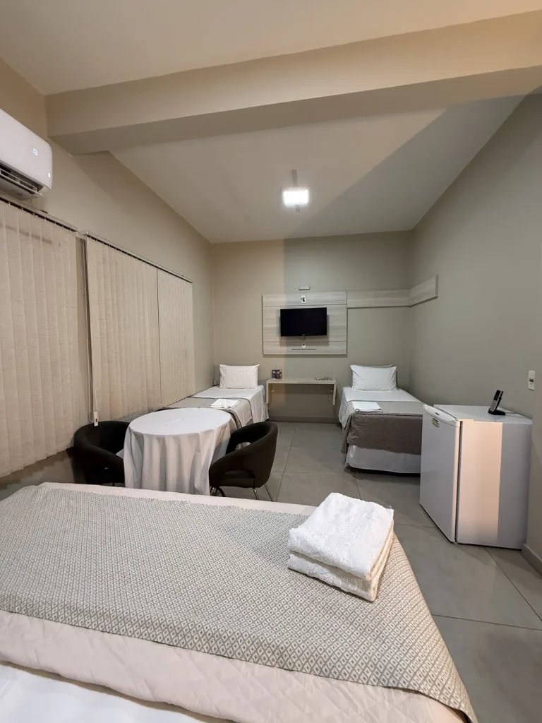 O quarto visto da cama queen, com mesa redonda, duas camas de solteiro, TV e frigobar