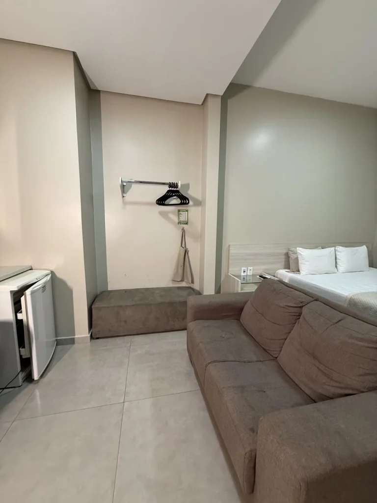 Cama queen ao lado do sofá, com frigobar e puff junto à parede