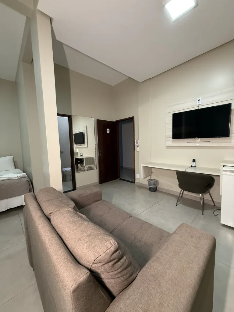 Sofá em primeiro plano, TV no painel e bancada de trabalho, com a porta do banheiro ao fundo