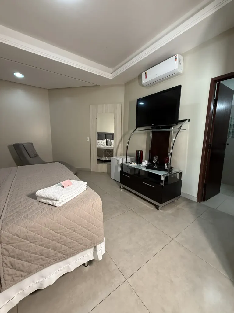 Cama queen em primeiro plano, com TV sobre rack, frigobar e porta do banheiro ao fundo