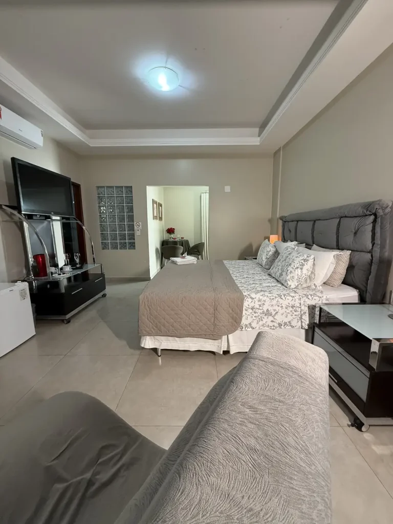 O quarto visto do fundo, com cama queen, TV, frigobar e parede de blocos de vidro