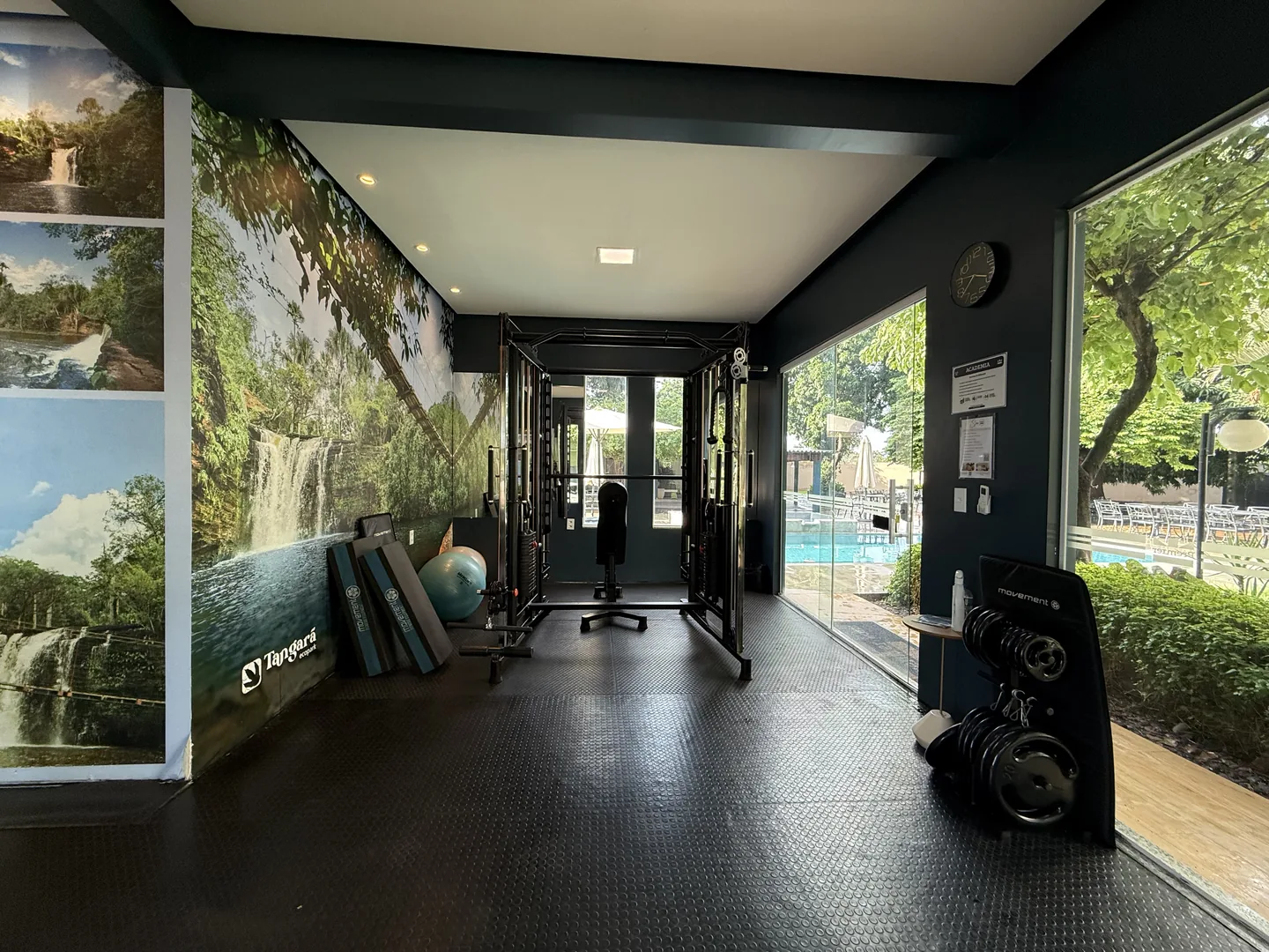 Academia vista da entrada, com painel de cachoeira na parede, estação de musculação ao fundo e janelas de vidro voltadas para a piscina