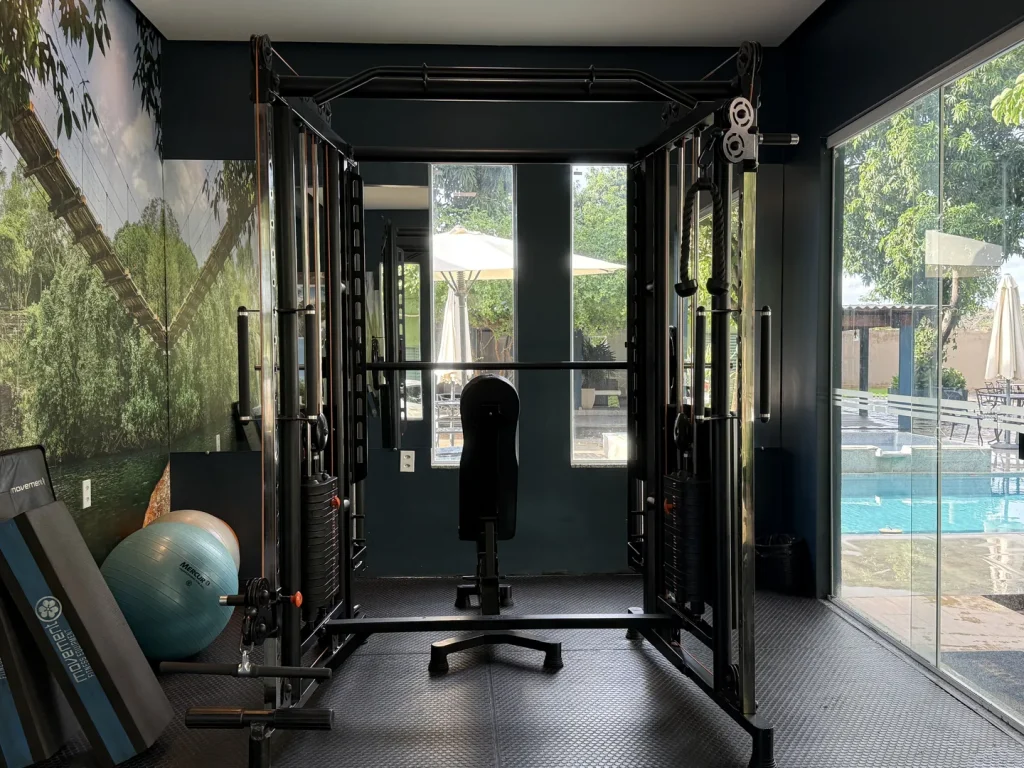 Estação de musculação com banco, bola de exercício e colchonetes, diante das janelas com vista para a piscina