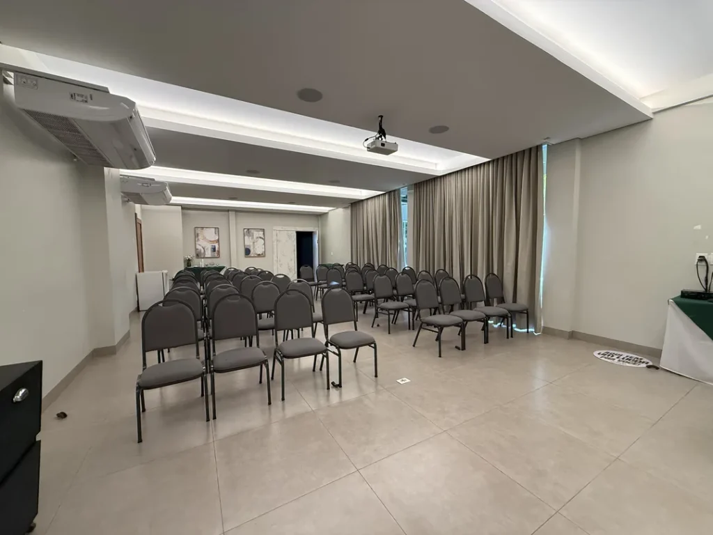Auditório Premier visto de lado, com fileiras de cadeiras, projetor no teto e cortinas bege