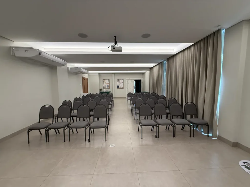 Auditório Premier visto do fundo pelo corredor central, com fileiras de cadeiras estofadas dos dois lados, projetor no teto e cortinas nas laterais