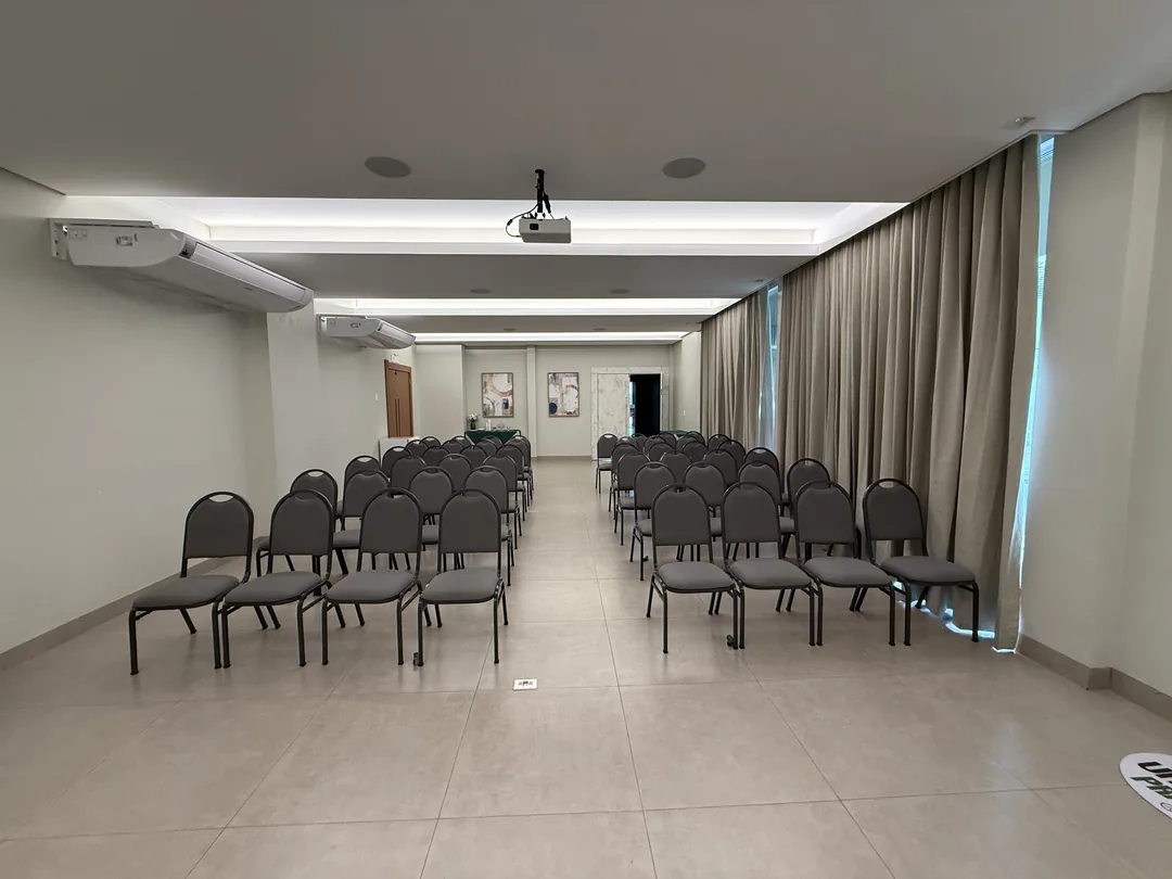Auditório Premier visto do fundo pelo corredor central, com fileiras de cadeiras estofadas dos dois lados, projetor no teto e cortinas nas laterais