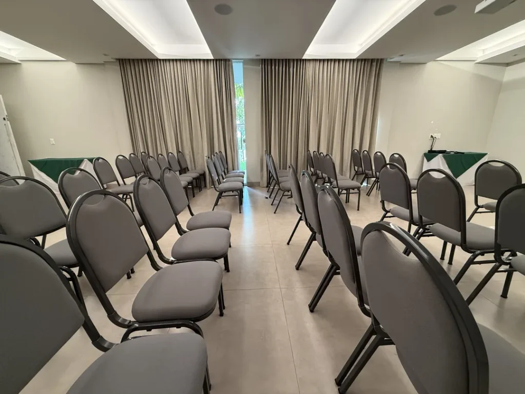 Fileiras de cadeiras estofadas cinza no Auditório Premier, com mesas de apoio de toalha verde ao fundo