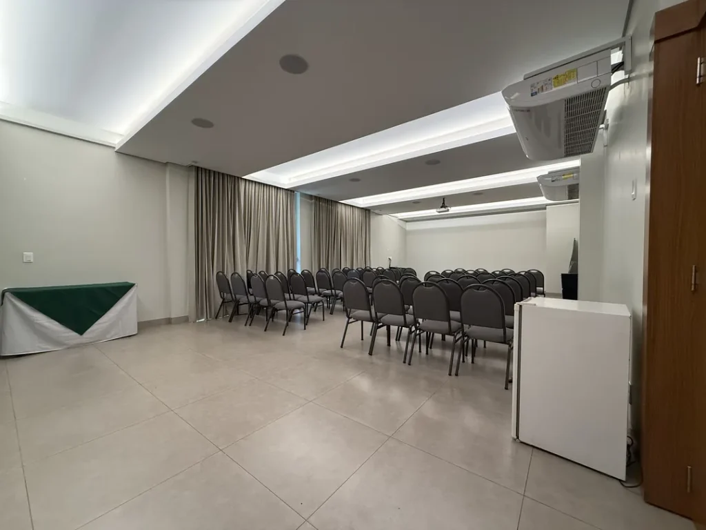 Vista da entrada do auditório, com mesa de apoio, fileiras de cadeiras e projetor no teto