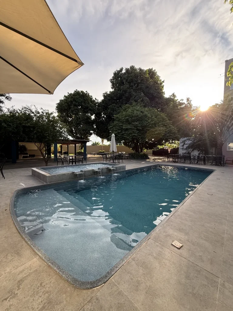Piscina ao entardecer, com o sol entre as árvores, cascata ao fundo e ombrelone em primeiro plano