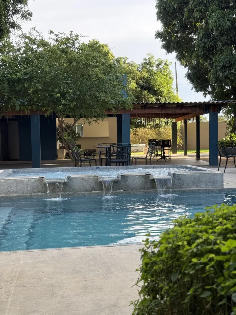 Cascata da piscina elevada caindo na piscina principal, com pergolado de pilares azuis e árvores ao fundo