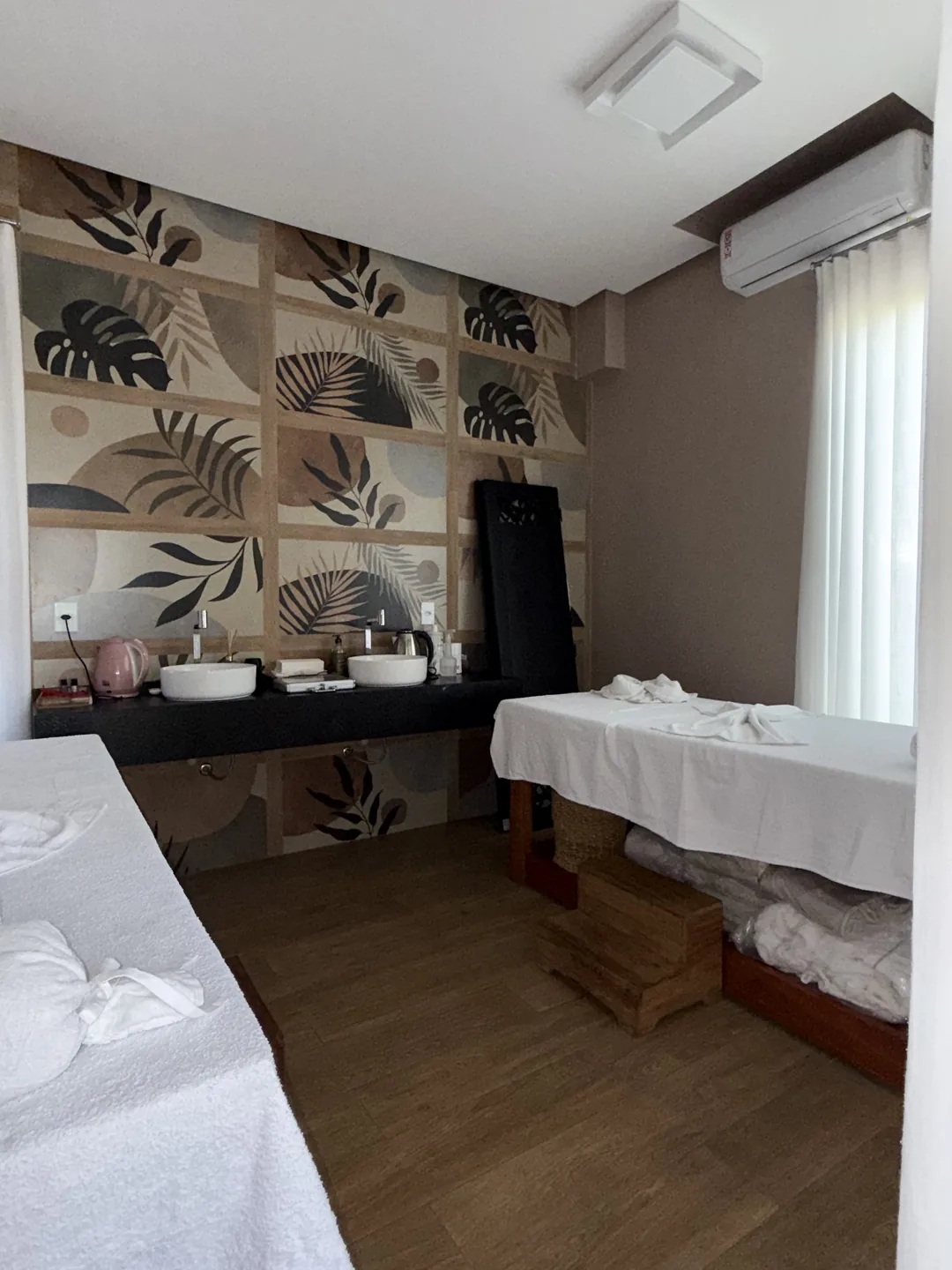 Sala de massagem com maca forrada de branco, bancada com duas cubas e painel de folhagens na parede