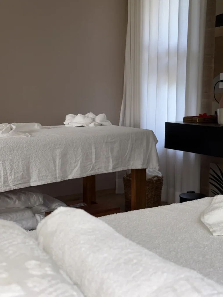 Detalhe de toalha enrolada sobre a maca, com outra maca e cortinas claras ao fundo
