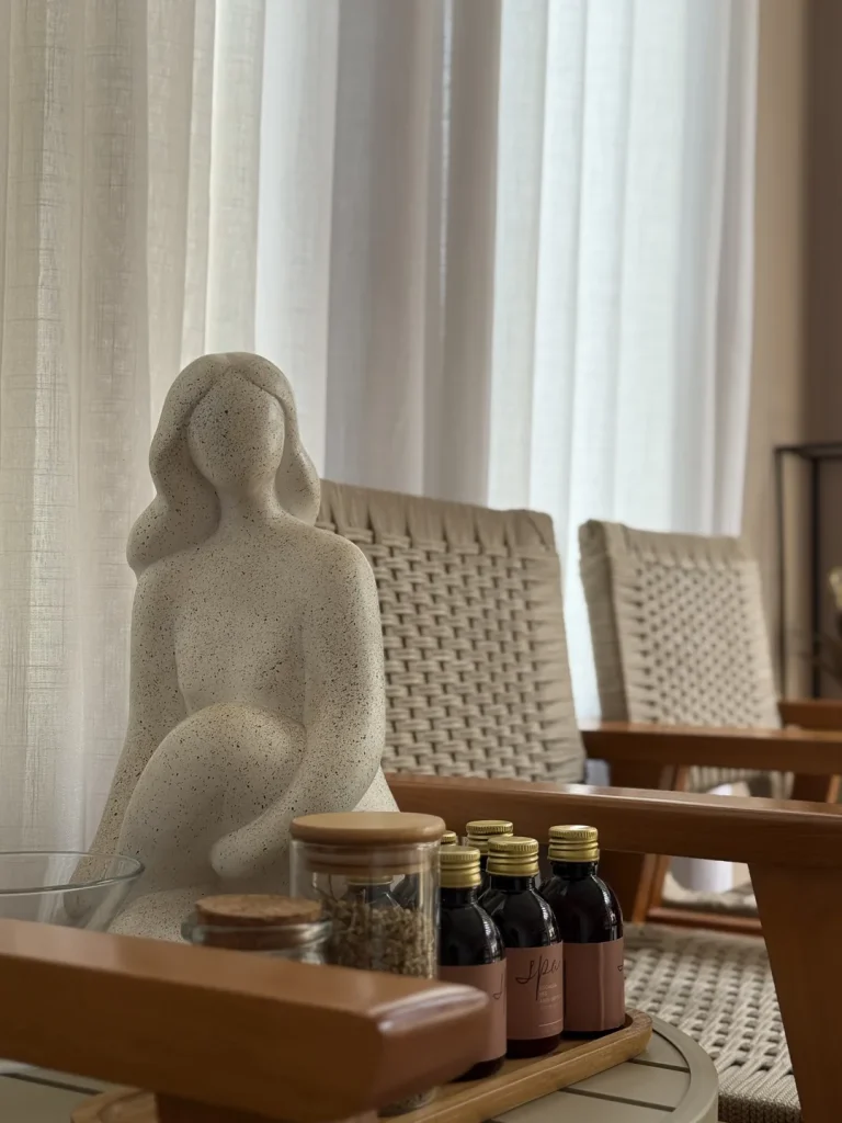 Escultura de figura sentada ao lado de frascos âmbar e pote de ervas, sobre mesa de apoio entre poltronas