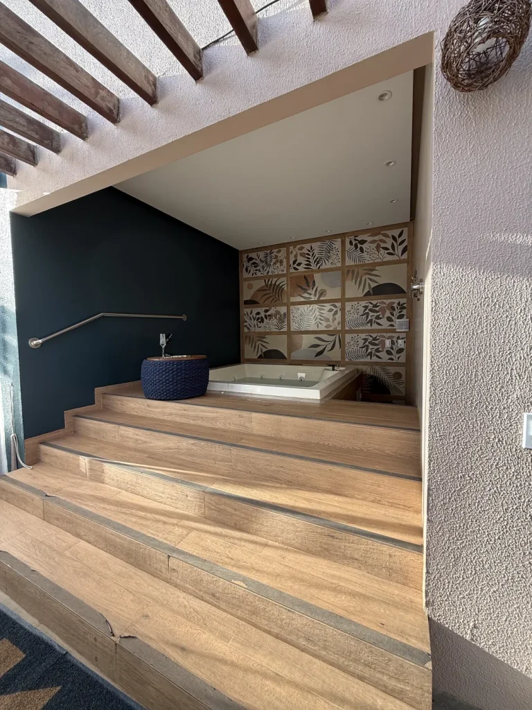 Banheira de hidro sobre plataforma de madeira com degraus, puff trançado azul e painel de folhagens