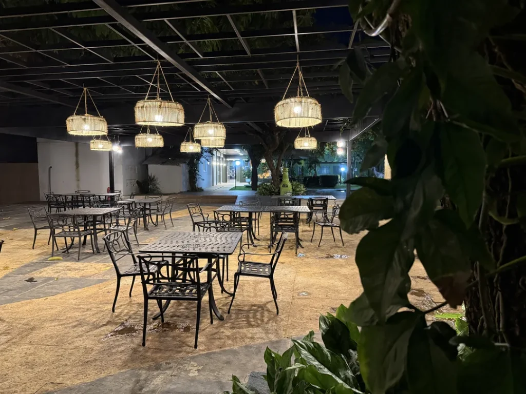 Mesas de ferro sob luminárias pendentes acesas, com plantas em primeiro plano