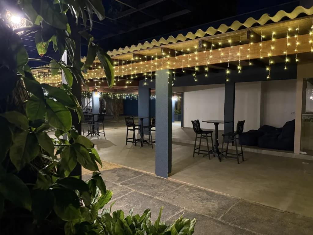 Pergolado com cordão de luzes e mesas altas, visto entre as plantas à noite