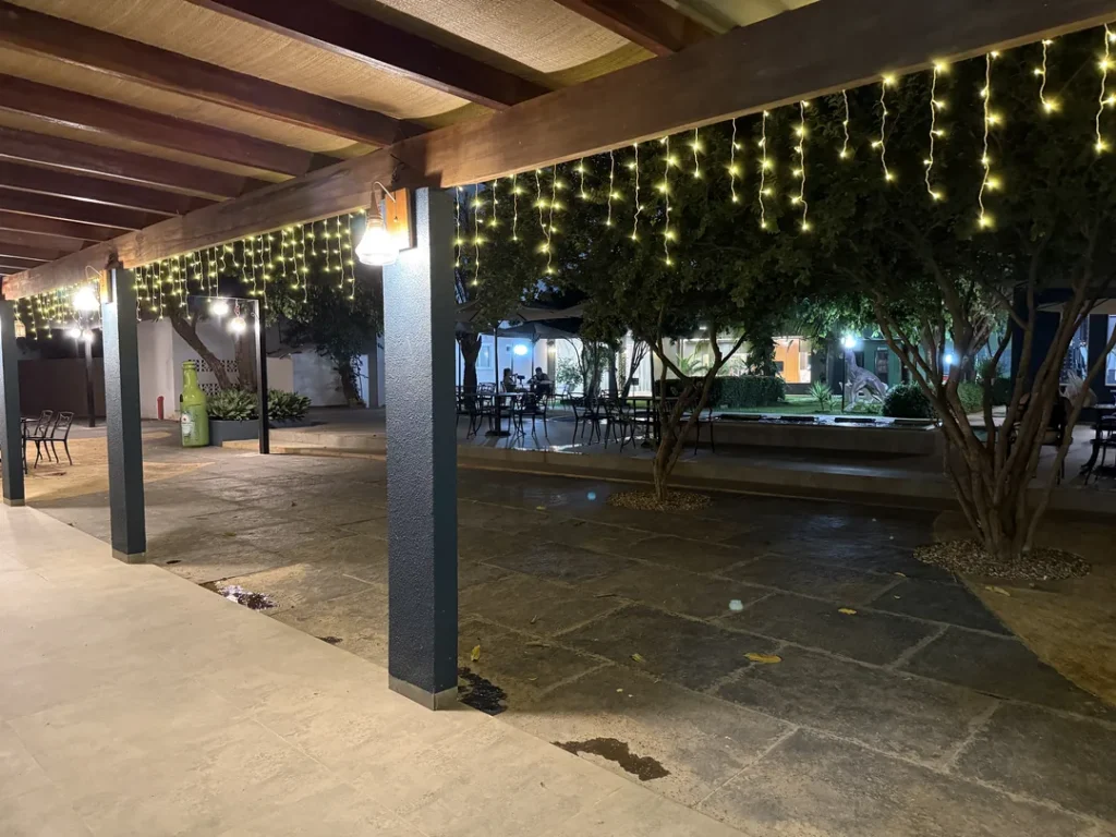 Área coberta com cortina de luzes, pilares azuis e árvores do jardim à noite